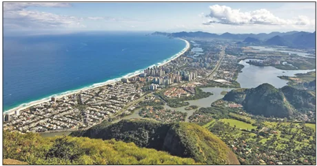 Fotografia wybrzeża Rio de Janeiro z góry: długa piaszczysta plaża nad oceanem, pas wieżowców, za nim laguny, a w tle i na pierwszym planie strome, zalesione wzgórza i masywy skalne.